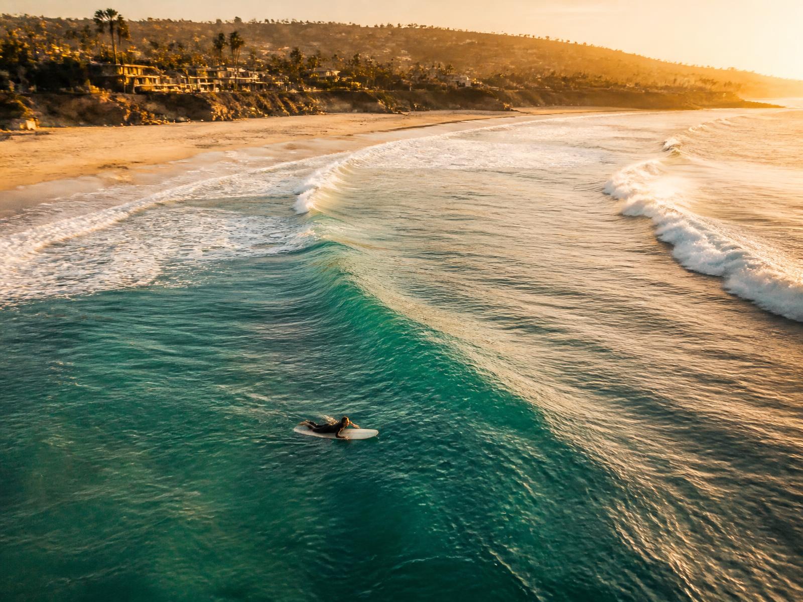 Aerial view of a surfer paddling out on a turquoise wave at golden hour